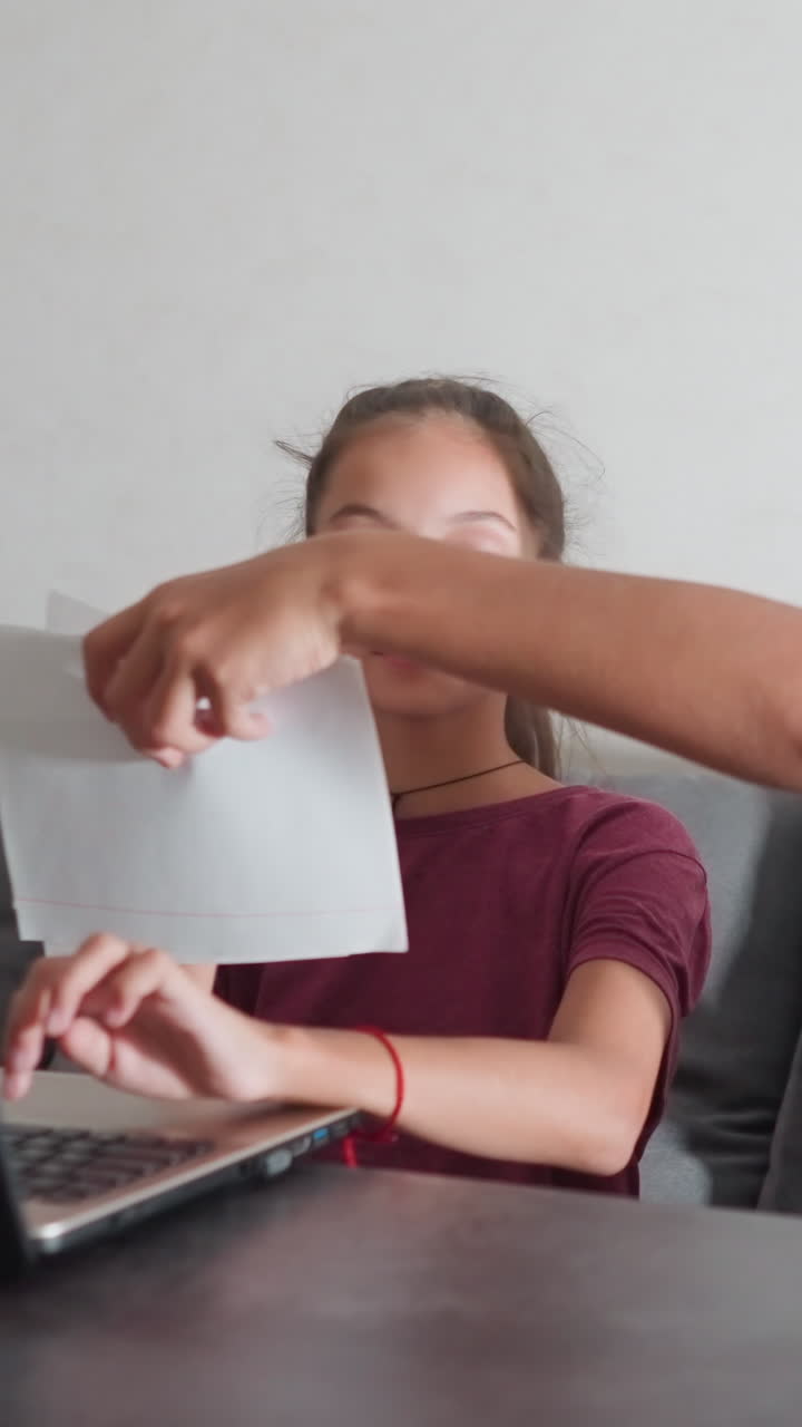 Una niña escribe en un portátil en el salón, distraída por su hermano, que está sentado a su lado mostrando los deberes que ha hecho. Ella echa un vistazo al trabajo de su hermano, sonríe y levanta el pulgar en señal de apoyo mientras estudian.