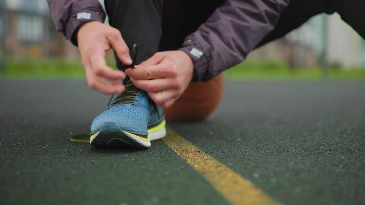 Atleta bianco si allaccia una scarpa da corsa blu, inginocchiato su un campo di asfalto vicino a una linea gialla, con un pallone da basket dietro. Le mani allacciano abilmente i lacci mentre le maniche della giacca si tirano indietro. Primo piano con ripresa dal basso.