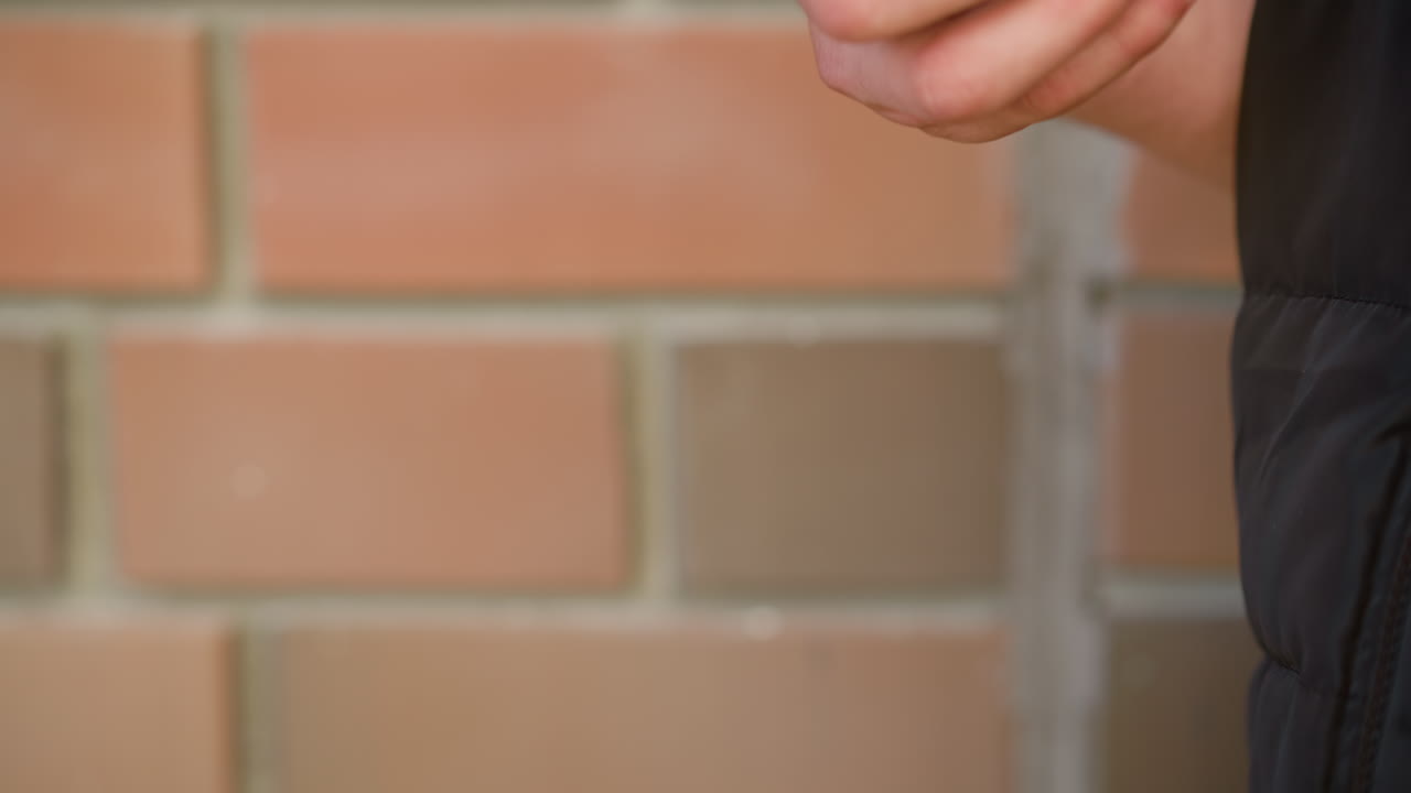 close up of white person holding vaping device in relaxed grip with fingers partially curled, standing beside softly blurred brick wall under daylight wearing dark puffer vest
