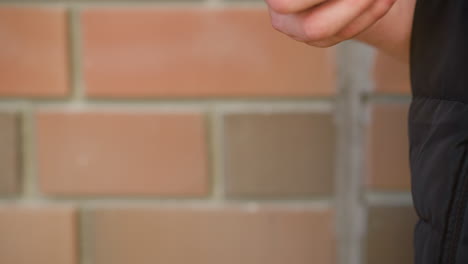 close up of white person holding vaping device in relaxed grip with fingers partially curled, standing beside softly blurred brick wall under daylight wearing dark puffer vest
