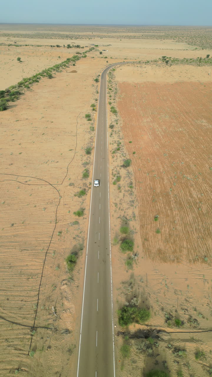 A forward-facing 4K vertical drone shot capturing a car driving on the Longewala Road on a sunny day with scattered bushesh Jaisalmer, Rajasthan.
