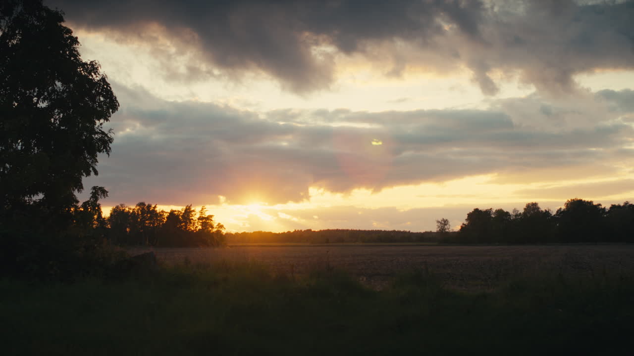 A serene rural landscape at sunset, with warm golden light spreading across open fields beneath dramatic clouds