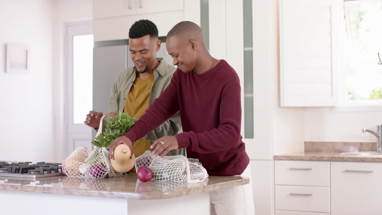 Happy african american gay male couple unpacking groceries in kitchen, slow motion