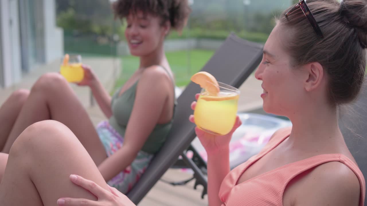 Ulike venninner snur seg, smiler på bassengkanten i badetøy, med drinker, deler en telefon og ler.