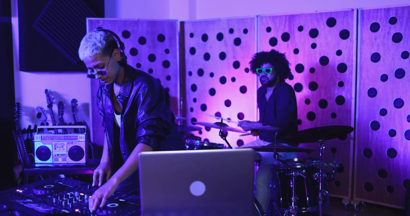 DJ che mixa musica con un batterista che suona in uno studio illuminato al neon.