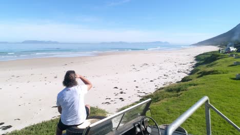 Drohnenaufnahme-Eines-Mannes,-Der-Mit-Einem-Surfbrett-Auf-Seinem-Landrover-Sitzt-Und-Auf-Das-Meer-Blickt,-Während-Die-Potenzielle-Brandung-Zum-Blick-Auf-Das-Meer-Wechselt