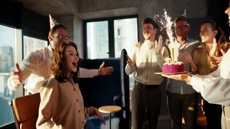 Colleagues-made-a-surprise-for-a-business-girl-in-a-brown-shirt-and-brought-her-a-cake-with-candles.-The-girl-is-very-happy-with