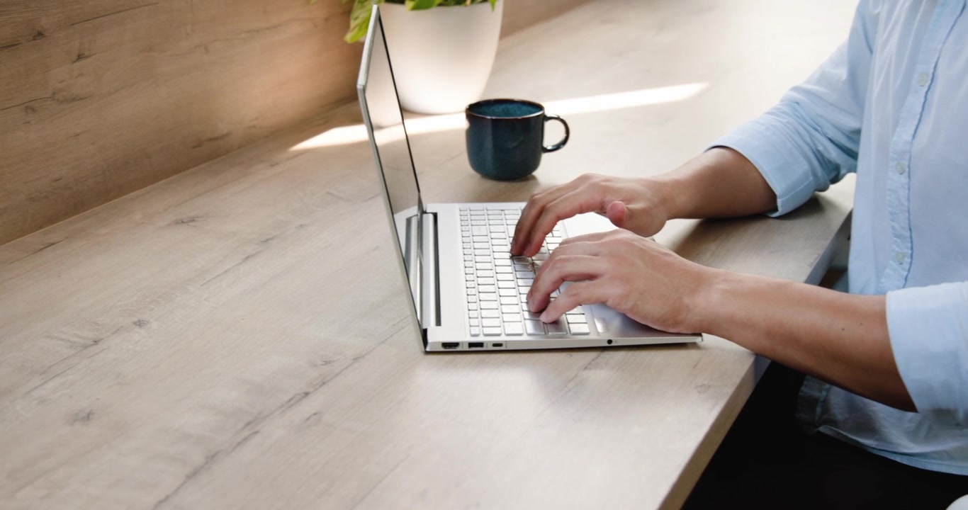 Asian man typing on laptop at desk, pausing, sipping from blue mug, resuming typing, copy space