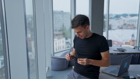 smiling dark-haired young man puts off his glasses. businessman standing in empty office thoughtfully looking through the window.