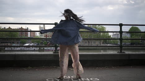 Fashionable-latina-tourist-with-black-wavy-hair-and-a-jean-jacket-walking-and-spinning-in-London-with-a-view-of-boats-and-buildings-behind-her