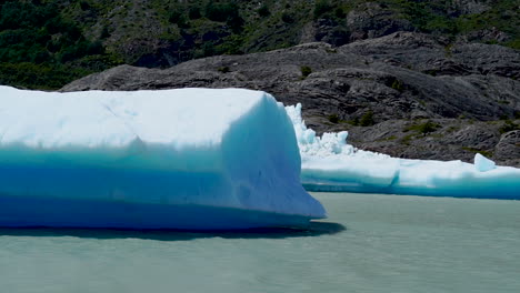 blue sunlit iceberg floating on grey lake, mountain in background