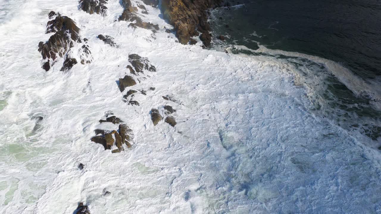 Drone footage captures powerful waves crashing against rocky coastline at Byron Bay, Australia, under bright daylight, showcasing nature's dynamic beauty