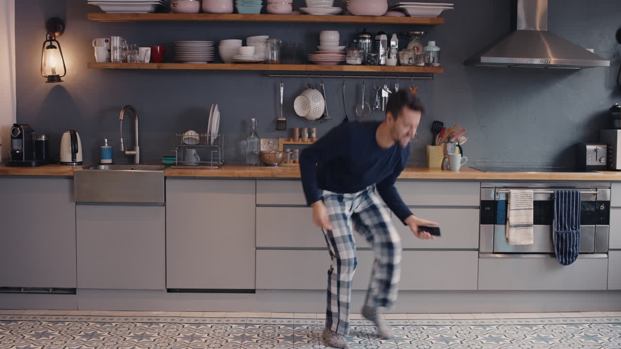 Happy young man dancing in kitchen wearing pajamas smart phone coffee morning