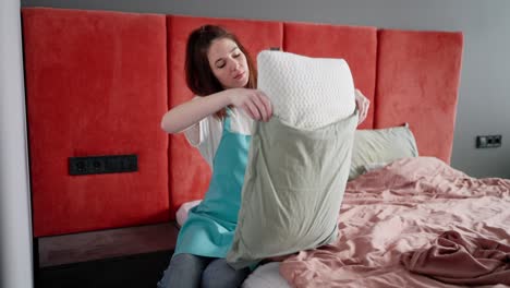 Side-view-of-a-brunette-cleaning-lady-girl-in-a-blue-apron-during-cleaning-tucking-a-white-pillow-into-a-gray-pillowcase-in-the-bedroom-when-ordering-a-cleaning-company-at-home