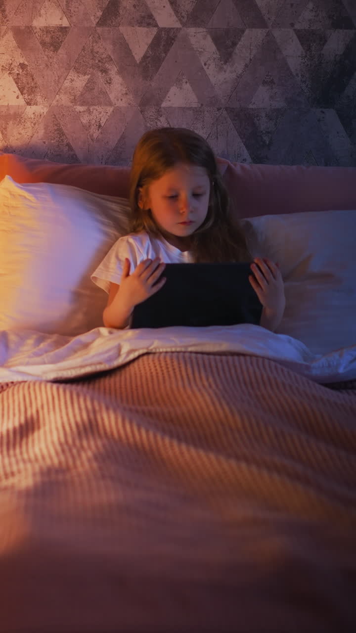 Cute little girl with long hair watches cartoons on tablet lying in comfortable bed by high decorative play tent with night lamp inside upper view slow motion