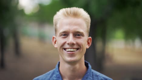 close up portrait young happy blond man standing in nature between park trees relaxes, breathes fresh air. male enjoys a life of peace calm, quiet in the park. outdoors. happiness