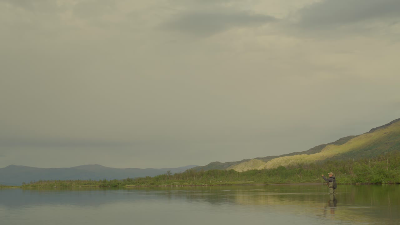 Man fly fishes in still lake in cloudy northern Sweden, wide side view