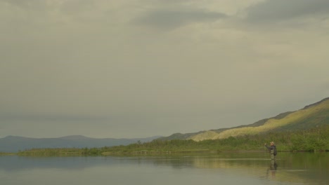 man fly fishes in still lake in cloudy northern sweden, wide side view