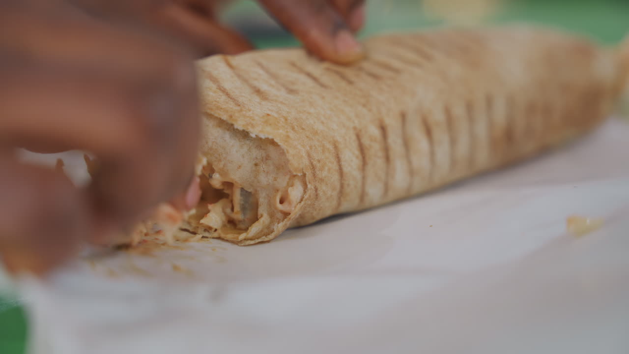Close-up tangan wanita kulit hitam membuka shawarma saat kencan piknik, roti pipih hangat, isian ayam empuk, saus krim, pembungkus kertas, remah-remah bertebaran, berbagi dengan lembut, cahaya alami yang lembut, meja kasual