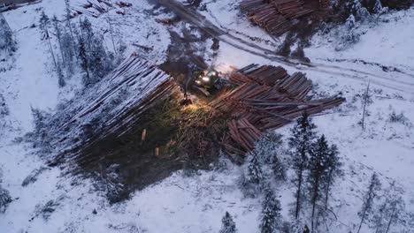 Die-Drohne-Bewegt-Sich-Dramatisch-Auf-Den-Holzmanipulator-Traktor-Zu,-Der-Im-Winter-Vor-Sonnenaufgang-Durch-Schneebedeckte-Haufen-Geschnittener,-Reifer-Kiefern-Im-Holzfällerlager-Sortiert