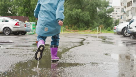 kid runs along wet parking site closeup active child in waterproof jacket and rubber boots plays on