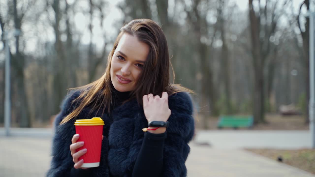 Beautiful woman with a plastic cup outdoors. Pretty girl in fur coat walking in the park. Back view of a brunette in the park. Slow motion.