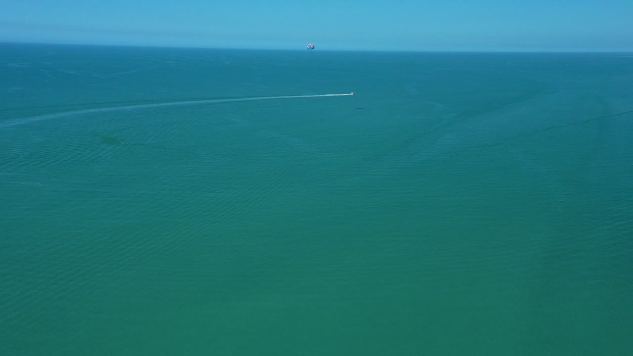 Una pequeña lancha motora se mueve en un amplio arco a través de un vasto y tranquilo océano cerca de una costa, dejando una estela blanca visible bajo cielos azules claros en un día soleado.