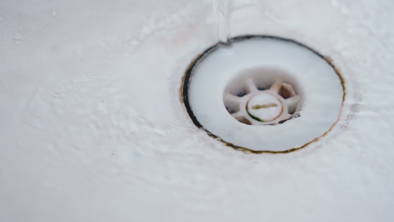 Gros plan sur un siphon de baignoire avec un anneau noir de taches et de saleté. La surface en porcelaine est inspectée, montrant des résidus de savon et une accumulation de calcaire autour du siphon. Un lent écoulement d'eau crée une ambiance de nettoyage domestique calme.