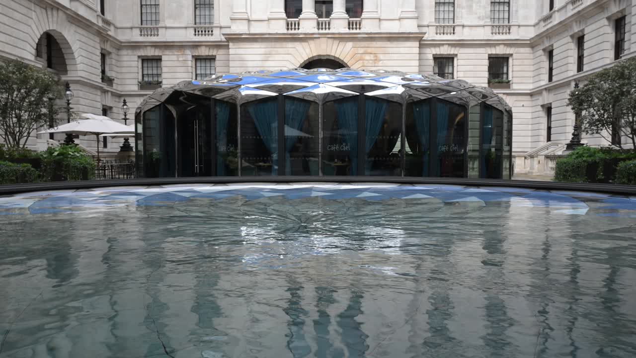 Una vista serena del exterior de una cafetería moderna reflejada en una instalación de agua estancada frente al histórico edificio Old War Office en un luminoso día de primavera.