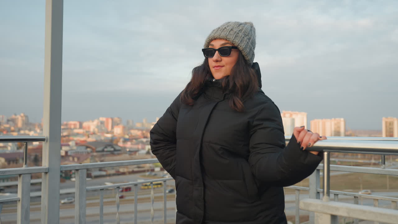 Donna che balla su un belvedere con sfondo di skyline, routine a corpo intero in lungo cappotto nero e cappello di lana, movimenti delle braccia deliberati e espressioni facciali giocose, occhiali da sole che fanno capolino, luce dorata fredda e casual