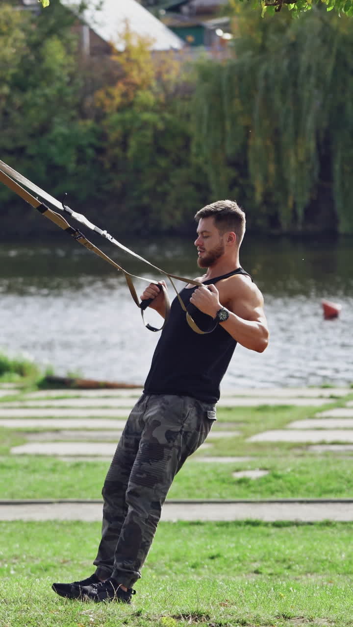 Man doing workout on trx near the river in summer. Young male exercising outdoors with straps. Side view of an athlete doing bungee training. Vertical video