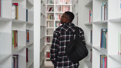 a young man with a backpack walks down an aisle in a library.