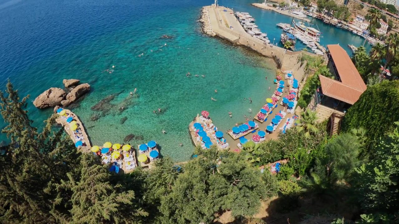 Veduta aerea della Marina di Kaleici con una baia mediterranea turchese, ombrelloni da spiaggia, nuotatori e storiche mura portuali ad Antalya, Turchia.