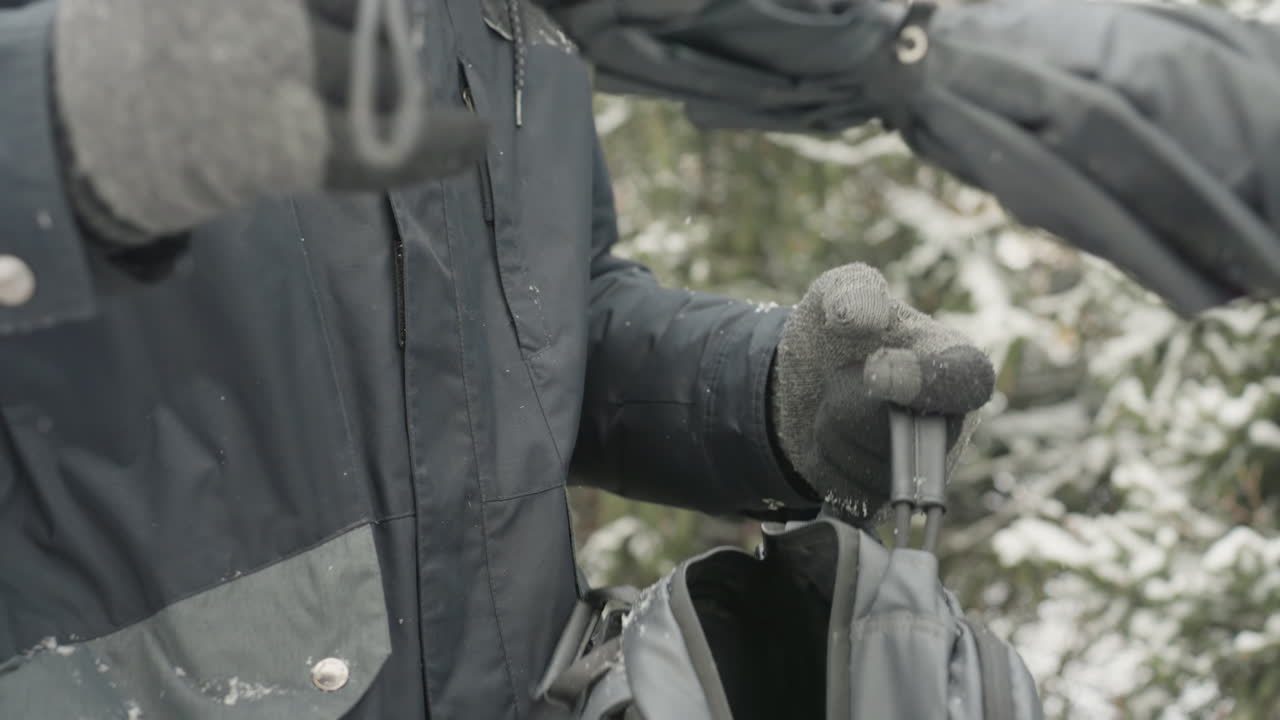 Hænder pakker rygsæk i sne, grå handsker og vandtæt jakke, lynlås lukkes, snefnug lægger sig på stof, forbereder kamera- og laptopudstyr, omhyggelig organisering og beskyttende lag.