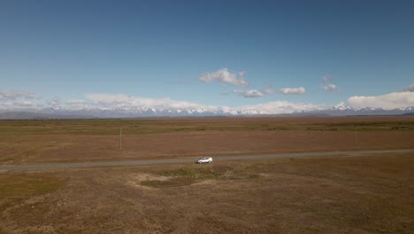 Empty-road-with-a-broken-down-car-within-a-flat-prairie-landscape,-mountains-in-background