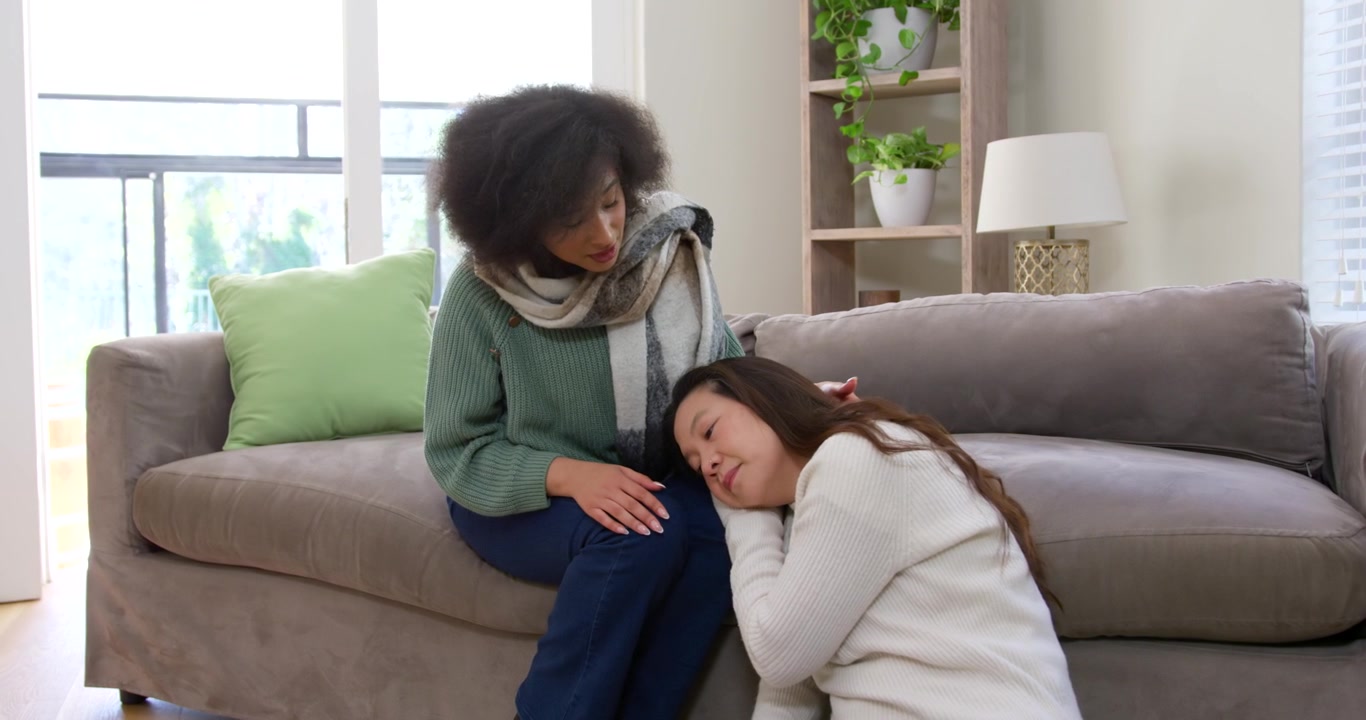 Diverse female friends: Asian woman seeking comfort, seated woman softly patting head on gray sofa