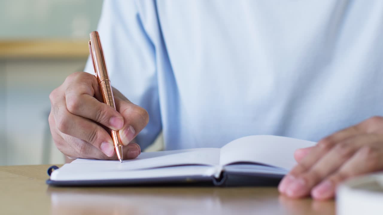 Man holding bronze pen hovering above left page, writing notes and steadying notebook for study