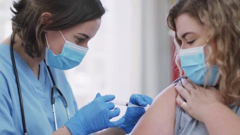 caucasian woman and caucasian female doctor wearing face mask at home vaccinating