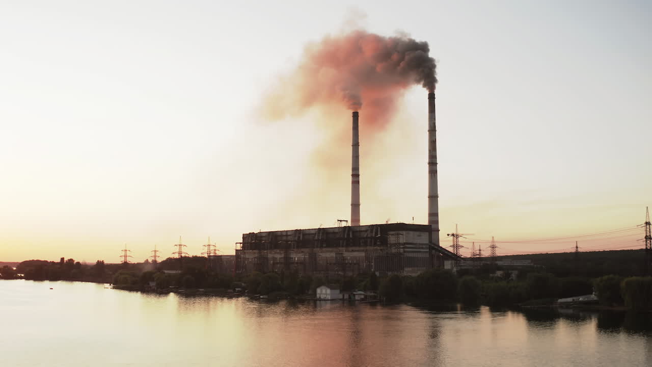Dark puffs of smoke releases into the atmosphere. Harmful emissions from industrial plant pollute the air near the river at sunset. Ecological disaster.