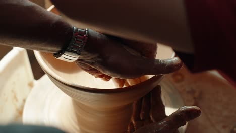 a pottery teacher and student, both men, shape a clay bowl together on a spinning wheel, their hands working in close collaboration during a lesson