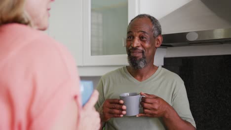 Over-shoulder-view-of-diverse-senior-couple-drinking-coffee-smiling-and-talking-in-kitchen