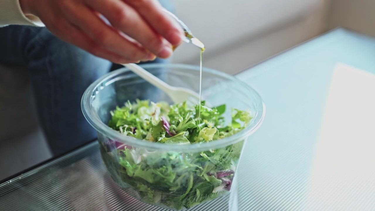 Person hælder dressing over frisk grøn salat i en plastikskål