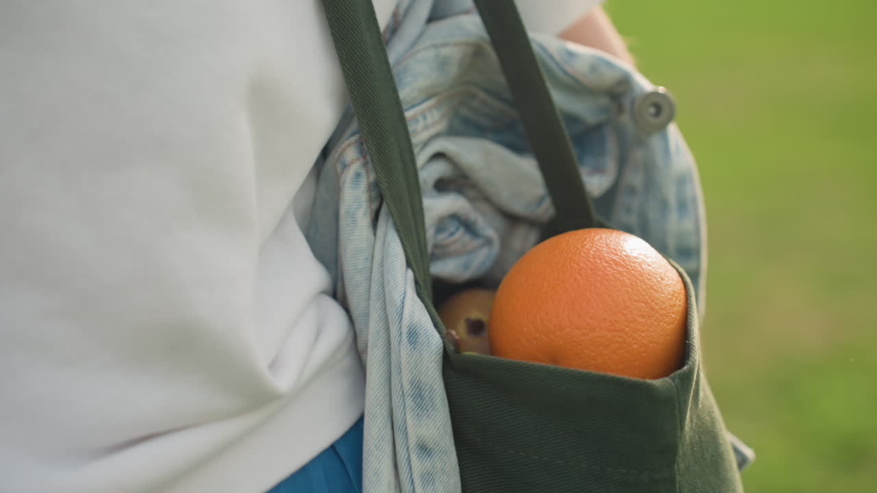 Close up side view of woman carrying canvas bag over shoulder holding fresh orange and denim fabric bright outdoor summer scene emphasizing healthy lifestyle and eco friendly shopping concept