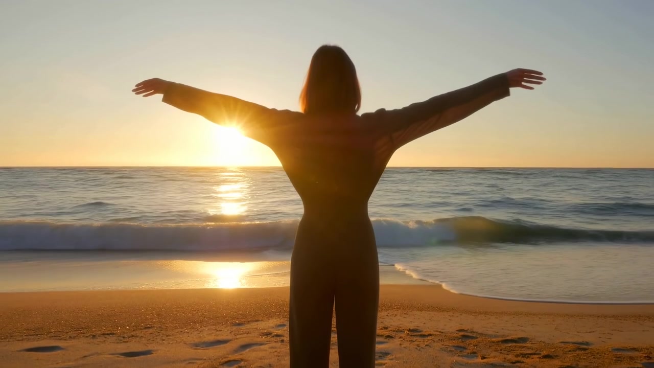 Silhueta de corpo inteiro de pessoa anônima em uma costa tranquila, de frente para o sol baixo com os braços levantados acima da cabeça em pose de celebração, cercada por um céu quente, ondas suaves, água reflexiva e luz suave do entardecer.
