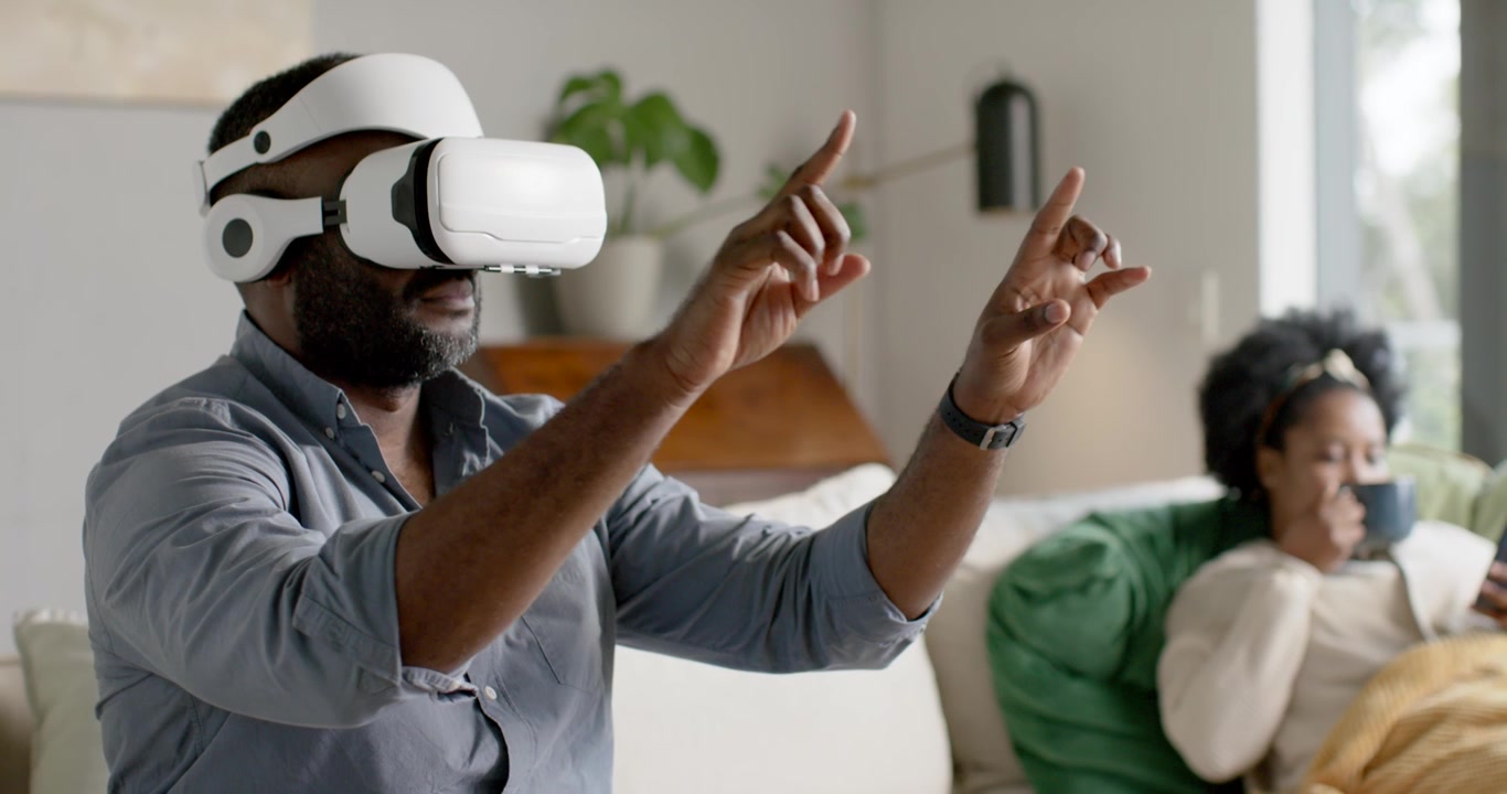 Afrikansk par på sofa, mand i headset gestikulerer mens VR starter; hun nipper til et krus og scroller på telefonen.