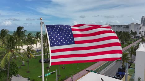an american flag waves near the sandy shores of south beach in miami, framed by palm trees and the atlantic ocean in the background. the scene looks towards the ocean from a beachfront park. aerial.