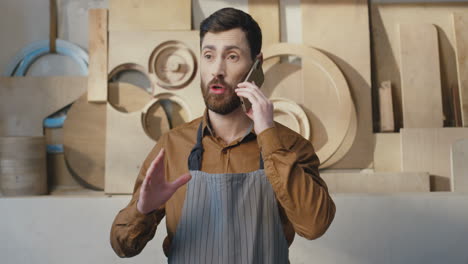 Portrait-of-caucasian-bearded-man-in-apron-talking-on-the-phone-in-carpentry-workshop