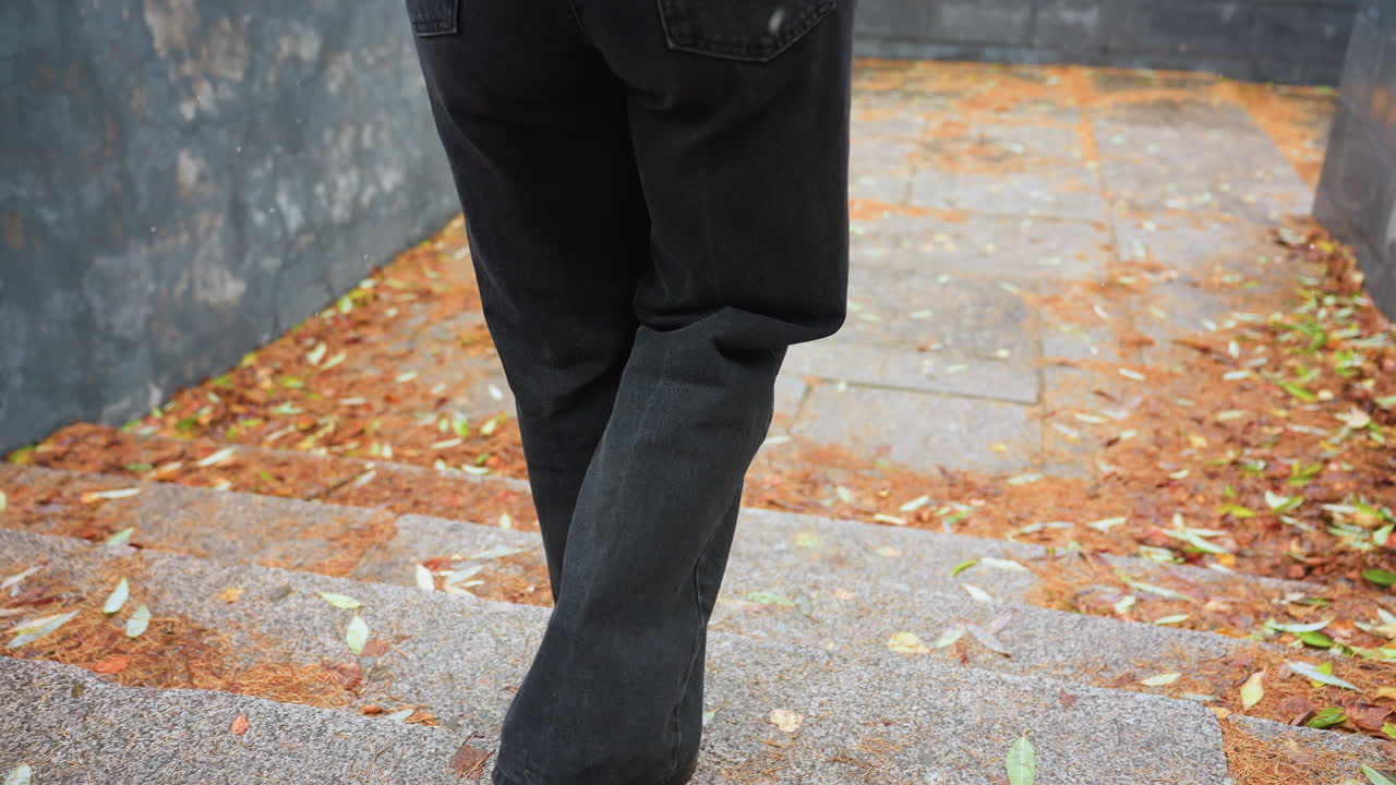 Legs of person walking down stone steps during light snowfall, wearing black jeans and black boots, colorful autumn leaves scattered along path, creating peaceful seasonal atmosphere