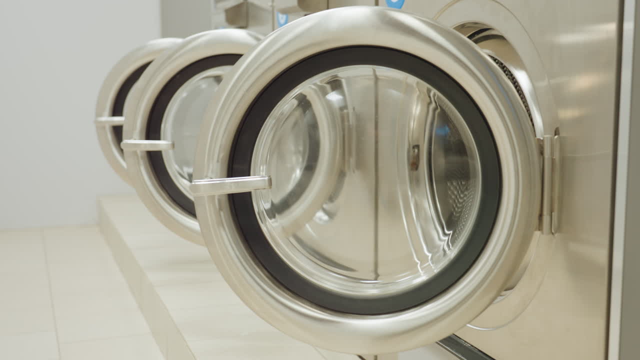 Dim room with industrial washers in a row, doors left open as faint blue control lights glow, environment gradually illuminated as light turns on, revealing metallic laundry equipment