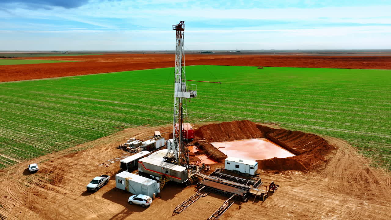 Green filed with oil industry production in the middle. Drone footage circling around the derrick with drilling equipment.